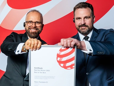 Two men in formal business attire—the left in a tuxedo with glasses, the right in a dark blue suit—jointly holding a framed award certificate with both hands. The certificate explicitly reads 