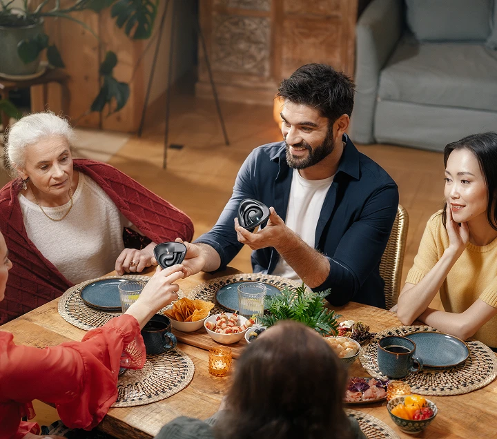 A diverse group of friends gathered around a table, sharing a meal and enjoying lively conversation, creating a warm, welcoming atmosphere as they pass a Vasco Translator E1 earbud to one another.