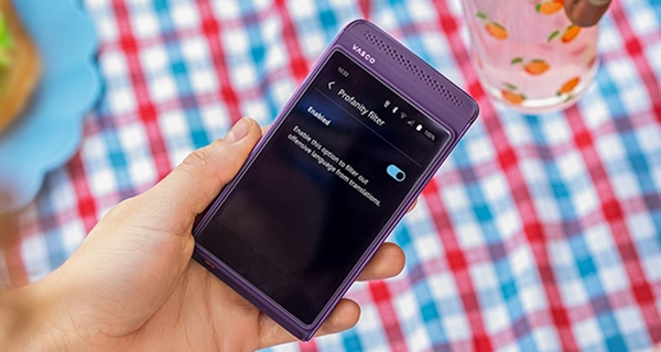 A close-up of a hand holding a purple Vasco electronic device. The display shows the "Profanity filter" settings menu, where the feature is "Enabled" with a blue toggle switch turned on, alongside text reading, "Enable this option to filter out offensive language from translations." The background features a red, white, and blue checkered tablecloth, with another hand reaching towards a clear glass decorated with peach illustrations in the top right corner.