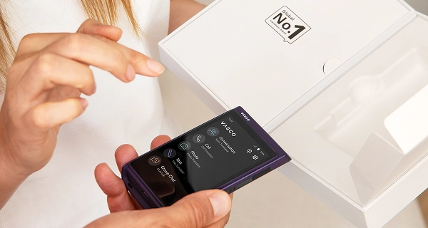 A close-up of two people interacting with a new device. One person is holding a dark purple pocket-sized electronic device, which displays a numeric dialling pad on its screen. Next to them, a woman with long brown hair, wearing a white blouse, reaches out to touch or point at the display. In the background, an open white product box is being held up, revealing a "No. 1" graphic logo on the inside of the lid.