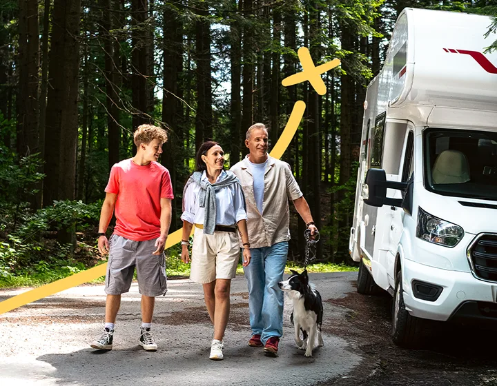 A family of three — two parents and one child — with a dog, walking away from their parked camper van on a sunny day. They are smiling, talking, and enjoying each other’s company.