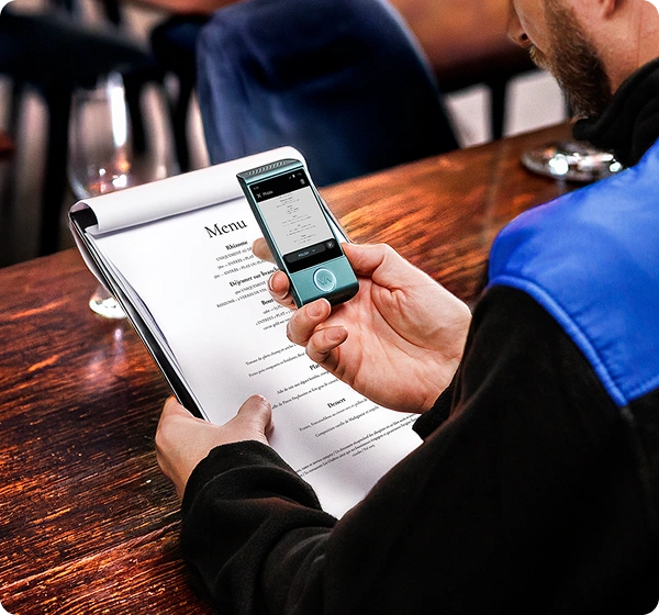 A close-up, high-angle shot of a person sitting at a wooden table in a restaurant, using a handheld electronic translator device to read a menu. The person holds a white paper menu in their left hand while holding a small, teal-colored Vasco translator over it with their right. The device’s screen shows a digital version of the menu being translated into Polish. The person is wearing a black jacket with a bright blue shoulder panel. The background is softly blurred, showing the warm interior of a dining establishment. 
